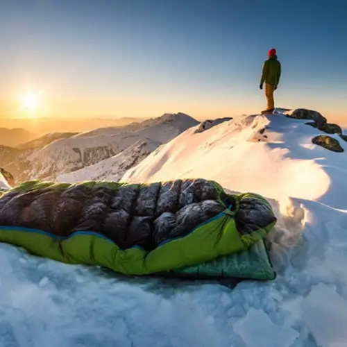 Homme au sommet d'une montagne en train de regarder le coucher du soleil à côté de son sac de couchage de survie.